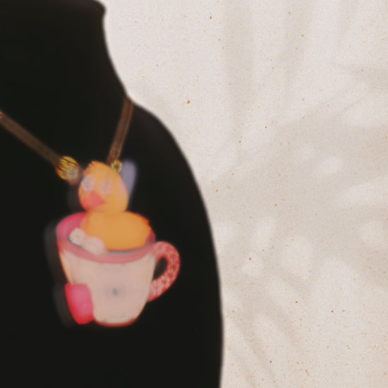 Video of the front and back of a mixed material handmade necklace of chibi cartoon duck sat in a pearlescent witch's brew teacup, with a gold chain and black gift box with a yellow familiars collection gift sleeve on black bust