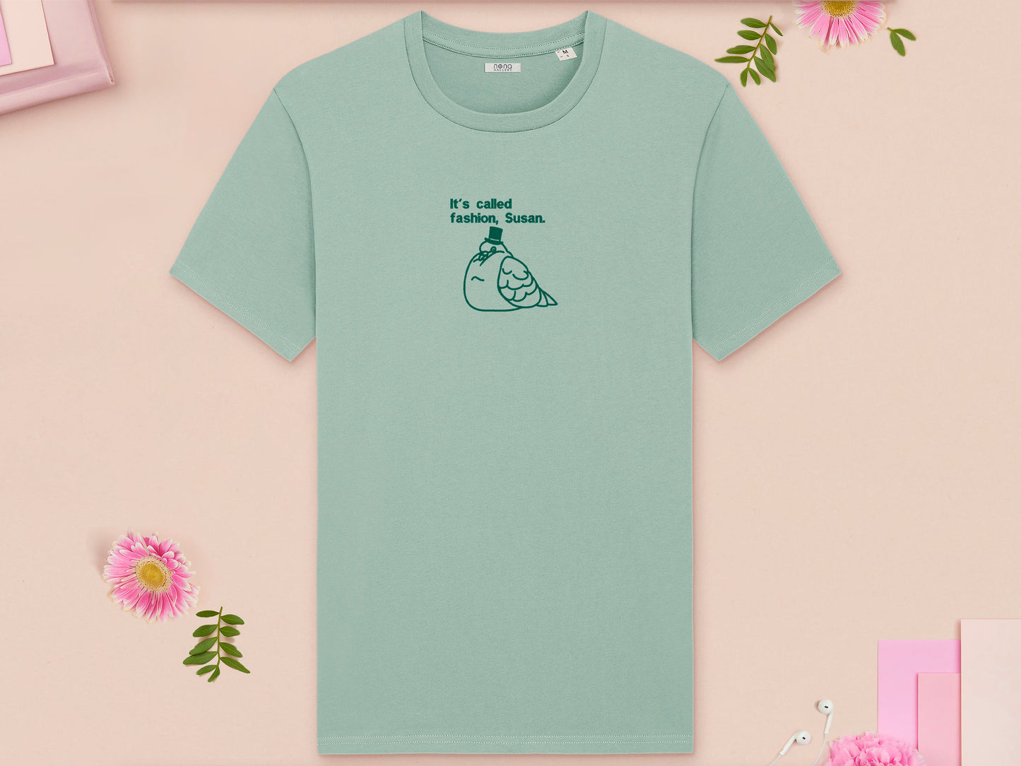 A green crew neck short sleeve t-shirt, with an embroidered green thread design of cute fat pigeon wearing a top hat with text underneath reading It's Called Fashion, Susan.