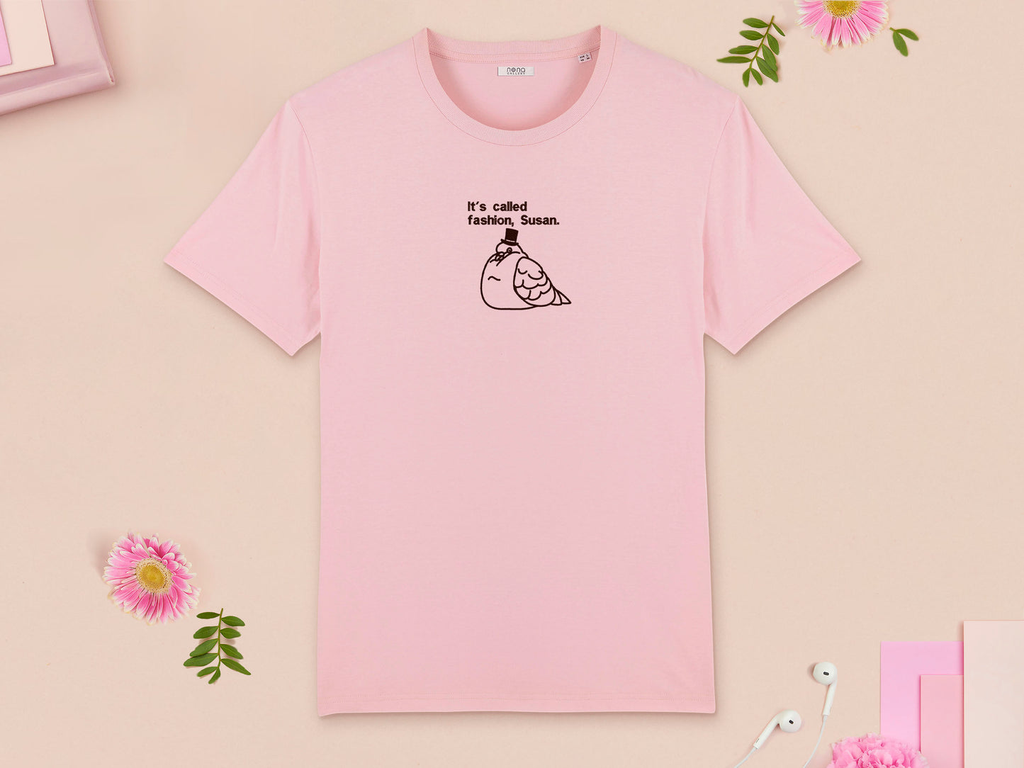A pink crew neck short sleeve t-shirt, with an embroidered brown thread design of cute fat pigeon wearing a top hat with text underneath reading It's Called Fashion, Susan.