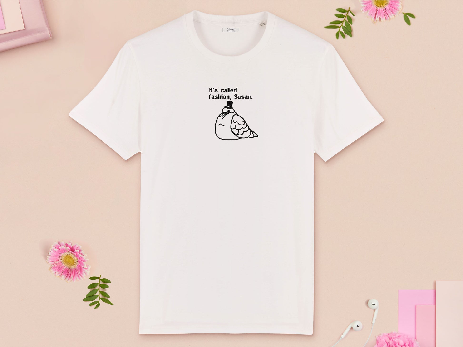 A white crew neck short sleeve t-shirt, with an embroidered black thread design of cute fat pigeon wearing a top hat with text underneath reading It's Called Fashion, Susan.